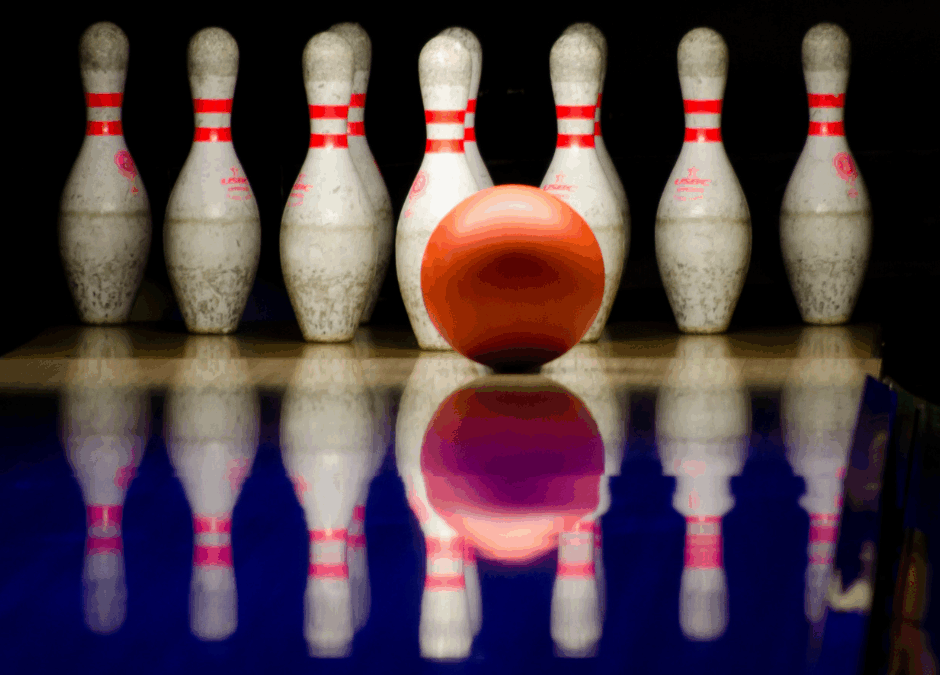A bright orange bowling ball rolls toward a set of seven white bowling pins with red stripes, reflected on the shiny blue surface of the bowling lane