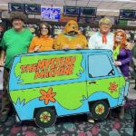 A group of people dressed as Scooby Doo characters pose with a large Mystery Machine van cutout at a bowling alley Bowling lanes and scoreboards are visible in the background