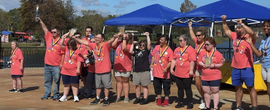 A group of people wearing matching pink shirts and medals stand outdoors with arms raised in celebration Some hold trophies, and blue tents and trees are visible in the background