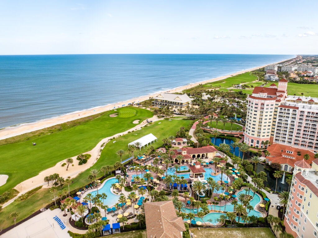 Aerial view of an oceanfront resort with multiple pools, palm trees, and lounge areas, next to a golf course and sandy beach, with the ocean and coastline stretching into the distance