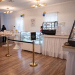 A bakery with glass display cases of pastries, a white counter, a red table, and a "Line Forms Here" sign Two people are behind the displays, and the room has wooden floors and decorated walls