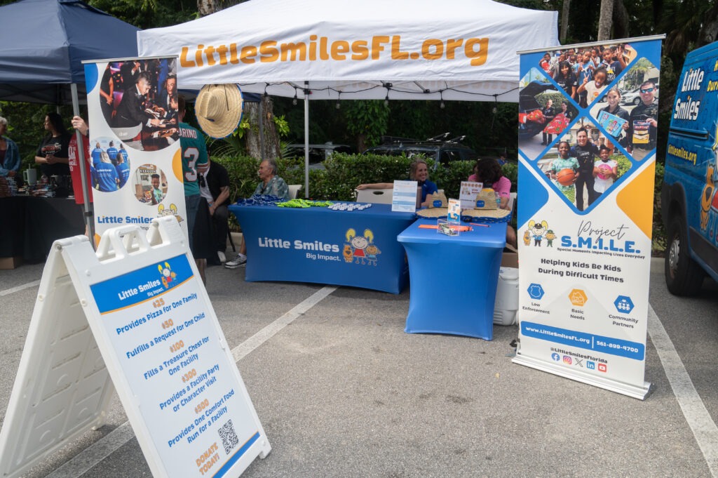 Outdoor booth for Little Smiles Florida with banners, an information board, and a table with volunteers and promotional items, promoting support for kids during difficult times