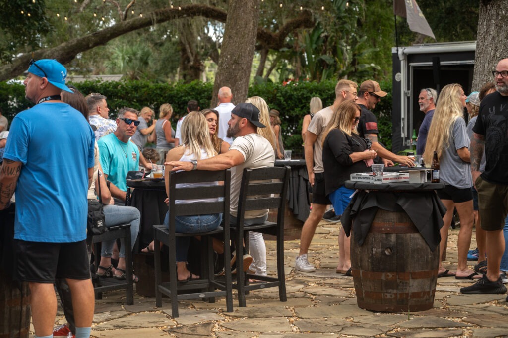 People gather outdoors at a casual event, sitting and standing around tables made from barrels Trees, string lights, and greenery create a relaxed atmosphere Most guests are chatting and enjoying food or drinks