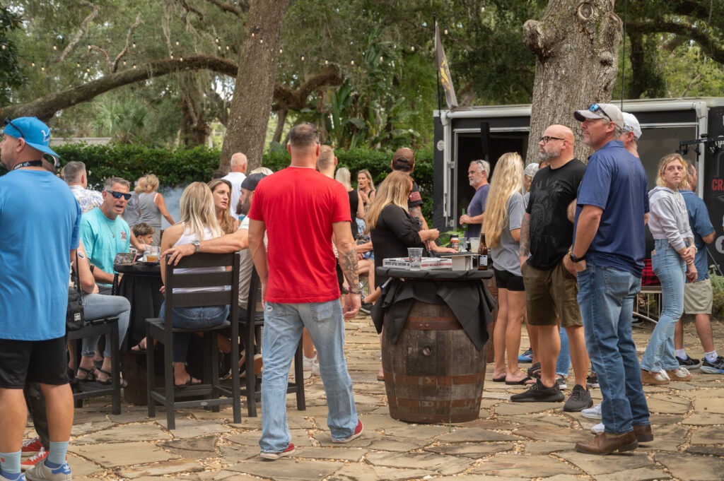 A group of people socialize outdoors on a stone patio under large trees, some standing and some sitting at tables Drinks and food are visible, and everyone appears relaxed and engaged in conversation