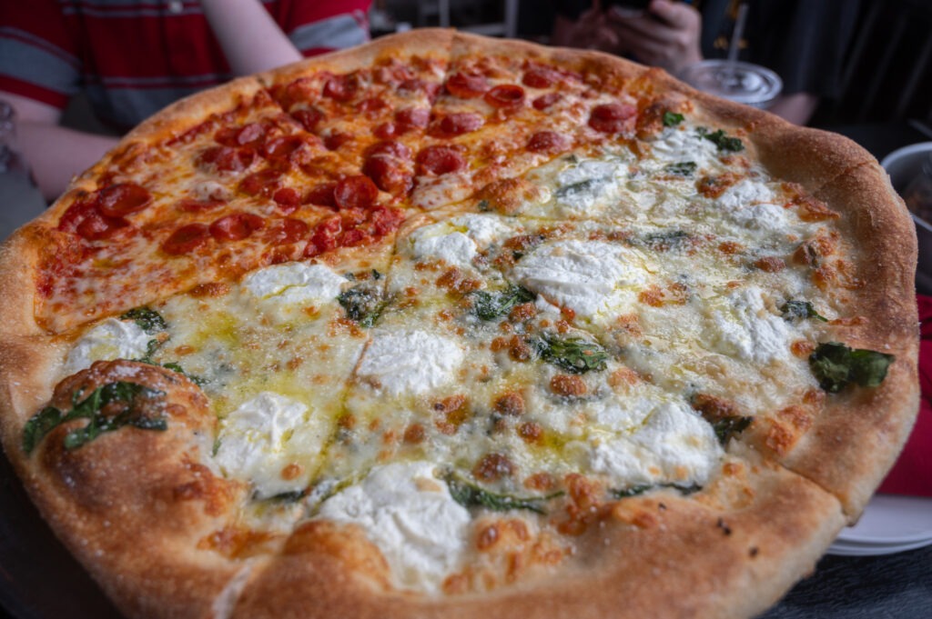 A large pizza with half topped with mini pepperoni slices and the other half topped with ricotta cheese and spinach The pizza sits on a table with drinks and people in the background