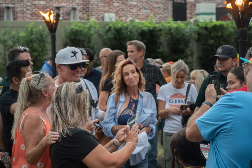 A group of people gather outdoors, some smiling and posing for photos A woman in the center holds a phone, while tiki torches burn nearby Trees and a brick wall are visible in the background
