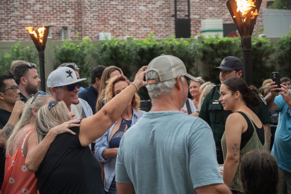 A group of people gathers outdoors near tiki torches, some smiling and posing for photos A woman in front is talking to the group while others take pictures Trees and a brick building are in the background