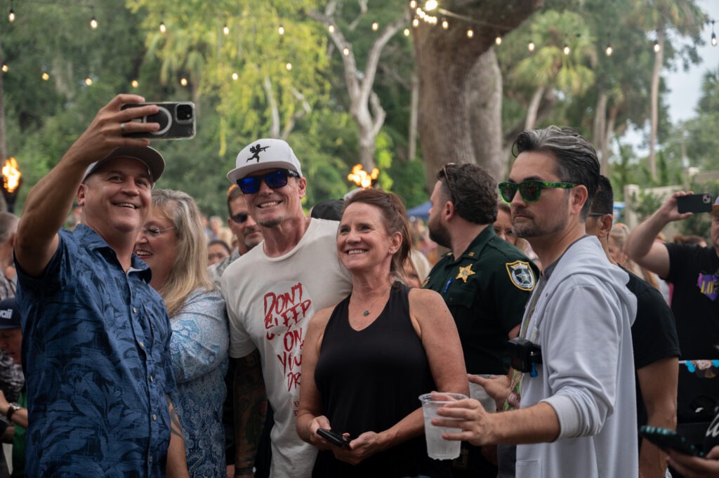 A group of people smiles and poses for a selfie at an outdoor event, with string lights and trees in the background One man holds a phone, and others wear sunglasses and casual clothing A police officer stands among them