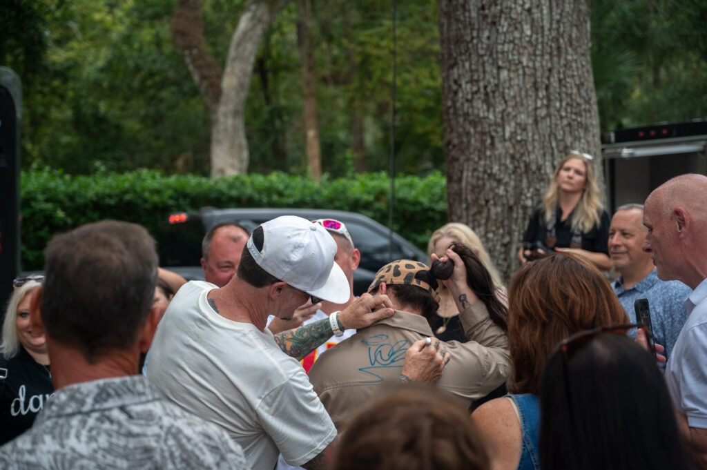 A man in a white cap signs the back of someone's shirt with a blue marker while a group of people watch outdoors, surrounded by trees and greenery
