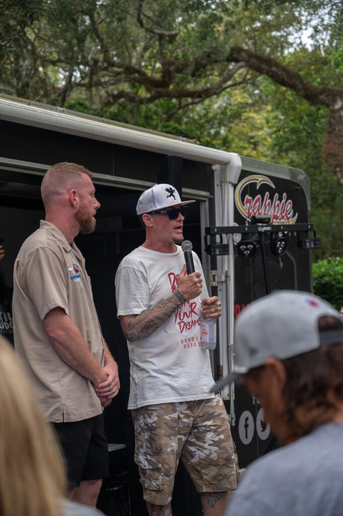 Two men stand and speak in front of a trailer with the "Crappie" logo; one holds a microphone and paper Trees are in the background, and an audience member wearing a cap is in the foreground