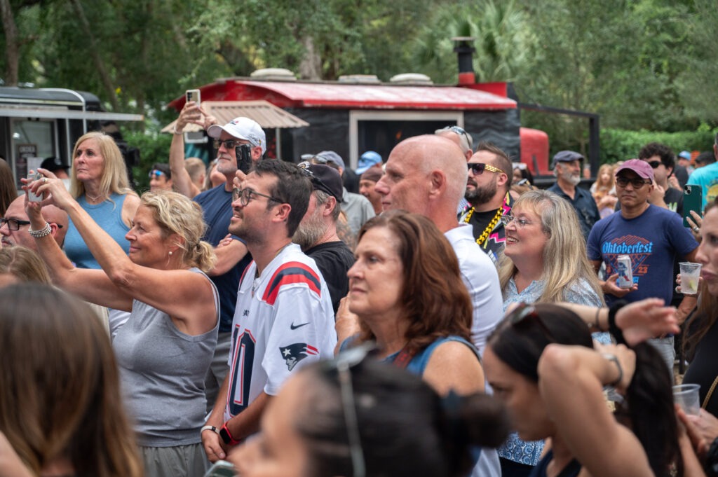 A diverse crowd of people, some smiling and taking photos, gather outdoors at an event near food trucks and trees on a sunny day
