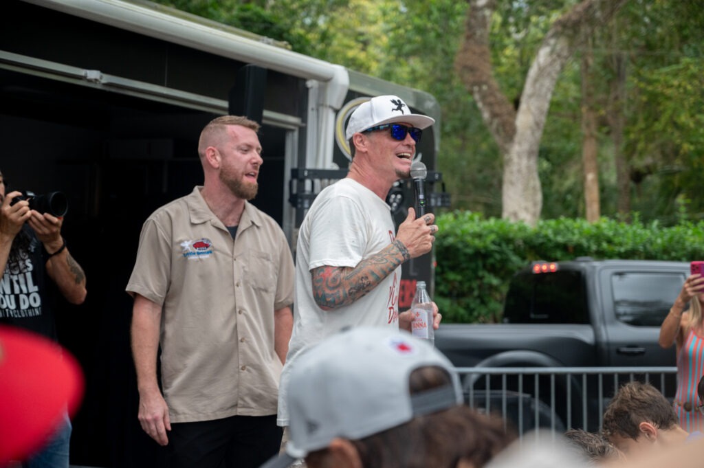 A man in a white t shirt, sunglasses, and a cap speaks into a microphone outdoors, while another man in a beige shirt stands beside him A crowd watches, and trees and a black truck are in the background