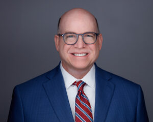 A smiling middle aged man with glasses, wearing a blue suit, white shirt, and red striped tie, poses in front of a plain gray background