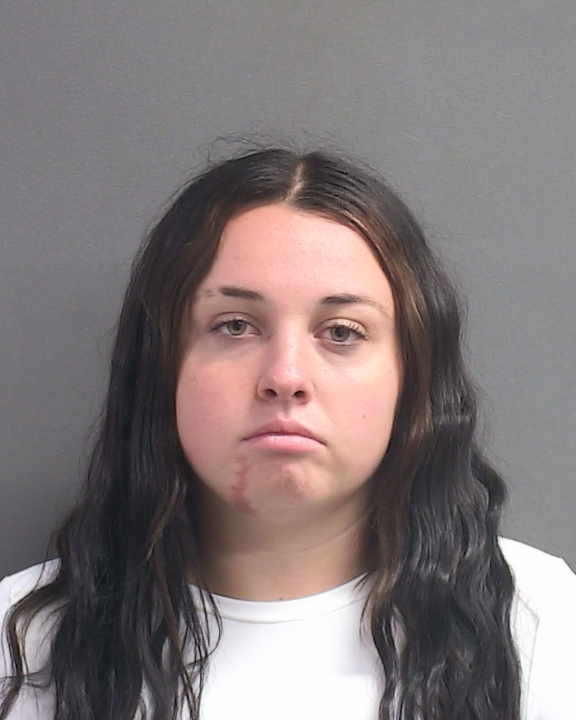 A young woman with long, wavy dark hair and a neutral expression stands against a plain gray background, wearing a white shirt