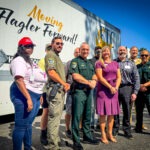 A group of people, including law enforcement officers and civilians, stand smiling in front of a truck with “Moving Flagler Forward!” written on the side, posing for a photo on a sunny day