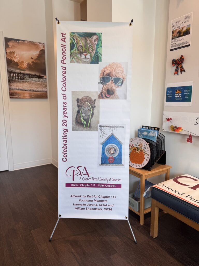 A vertical banner displays animal portraits and text: "Celebrating years of Colored Pencil Art" with the Colored Pencil Society of America logo and credits to artists The banner stands in a room with art and furniture