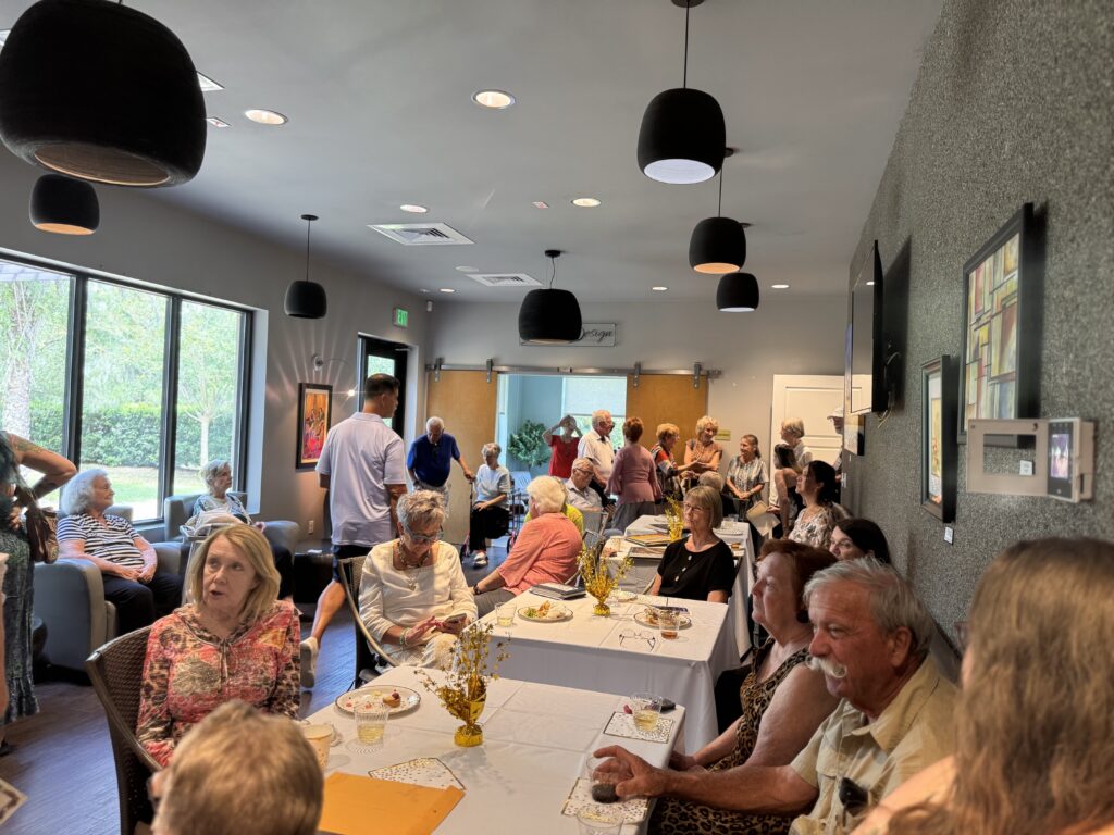 A group of people, mostly seniors, are gathered in a bright, modern room with large windows, sitting and standing around tables set with food and drinks, engaging in conversations and enjoying a social event