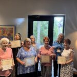 Seven smiling women stand indoors, each holding a certificate They are dressed in colorful tops and are positioned in front of a door and artwork The atmosphere appears joyful and celebratory