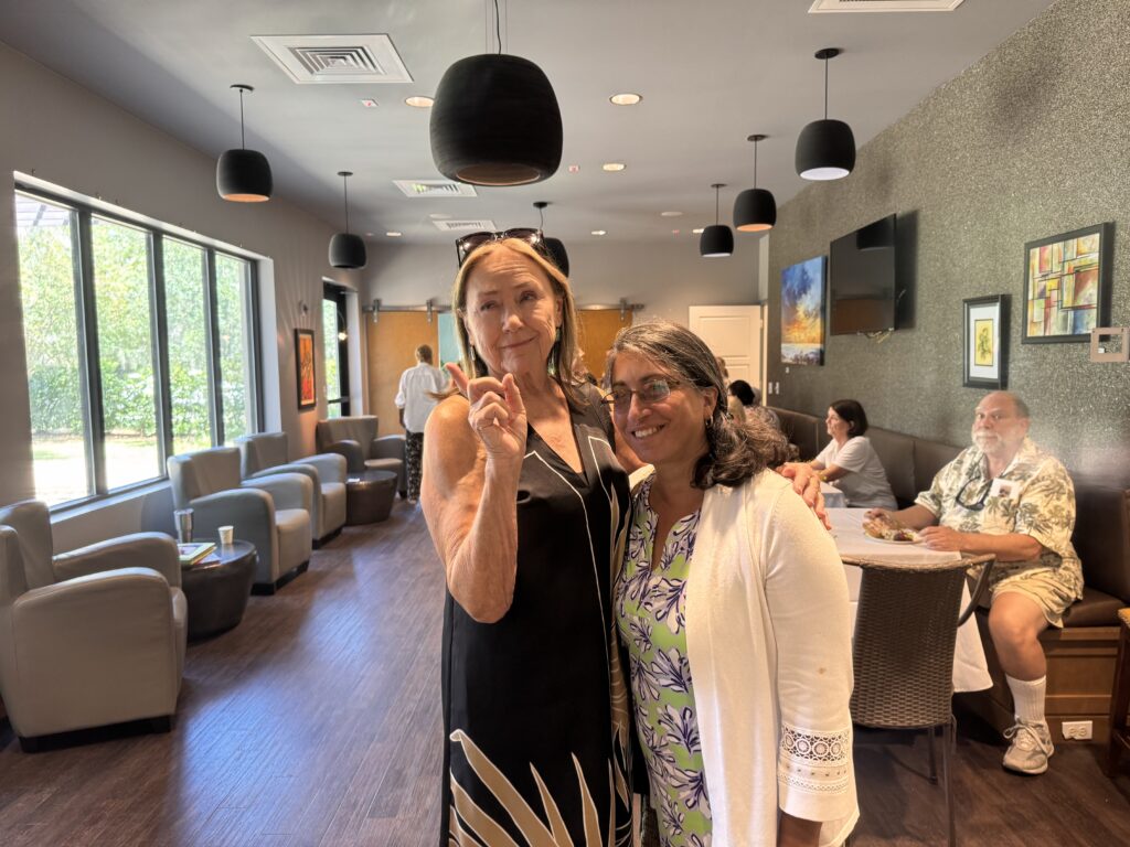 Two women stand together smiling in a bright lounge with modern decor, armchairs, and people sitting at tables in the background One woman gestures peace with her fingers while the other stands beside her, both looking at the camera