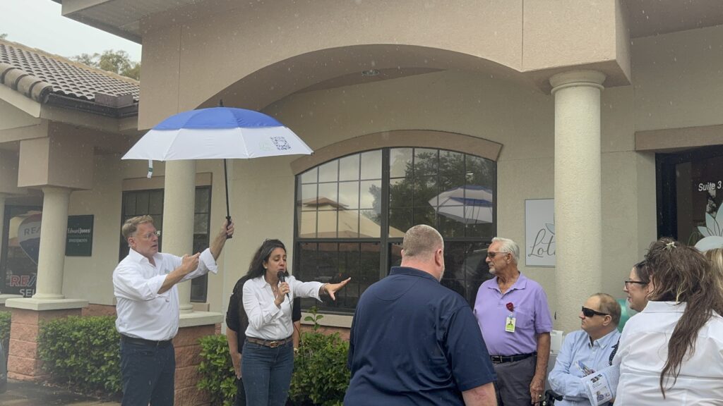 A woman speaks into a microphone to a small group outside a building A man holds a blue and white umbrella over her Several people listen, some wearing sunglasses, and the sky appears overcast or rainy