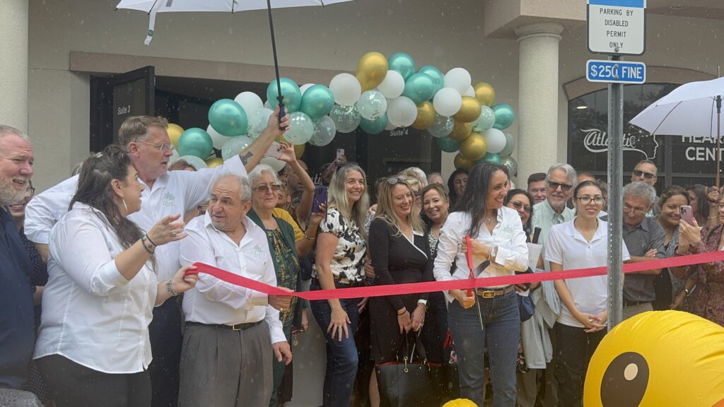 A group of people stands in front of a balloon arch at a ribbon cutting ceremony Some hold umbrellas in light rain, and a few are holding a large red ribbon There is a $ fine parking sign in the foreground