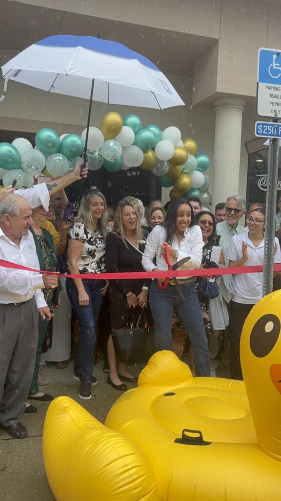 A woman cuts a red ribbon with large scissors at a ribbon cutting ceremony, surrounded by smiling people Balloons and an umbrella are overhead, and a large inflatable duck is in the foreground