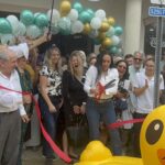 A woman cuts a red ribbon at an outdoor event, surrounded by smiling people, balloons, a large rubber duck float, and someone holding an umbrella A blue handicapped parking sign is visible on the right