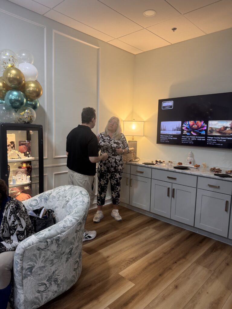 Two people stand and talk in a cozy room with balloons, a TV displaying various images, and a countertop with snacks Light wood flooring, white cabinets, and a soft armchair create a relaxed atmosphere