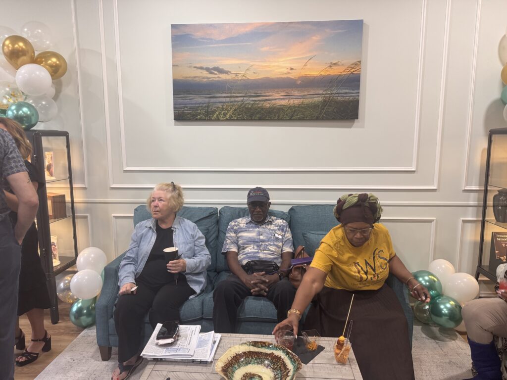 Three people sit on a blue couch in a decorated living room with balloons and a beach sunset painting on the wall One woman reaches for food, while the others relax, holding drinks