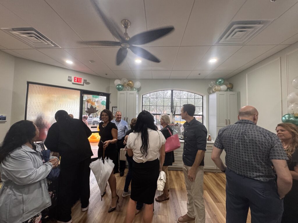 A group of people socialize in a decorated room with balloons, wood flooring, and large windows Most are standing and talking, while some hold bags or drinks Ceiling fan and light fixtures are visible above