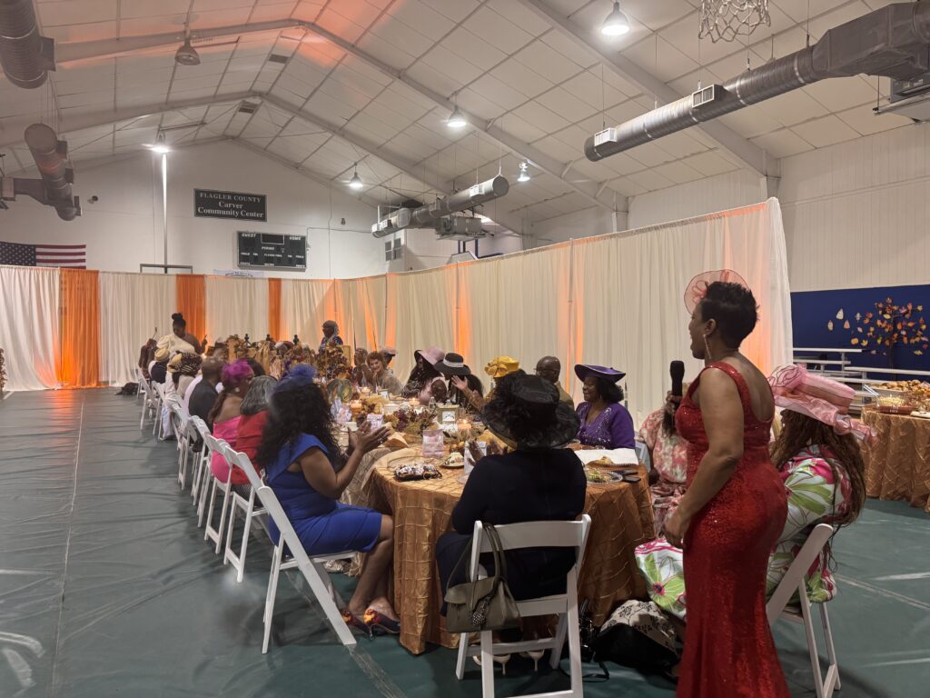 A large group of women in colorful dresses and hats are seated at long, decorated tables inside a gymnasium for a formal event One woman in a red dress stands and speaks, while others eat and converse