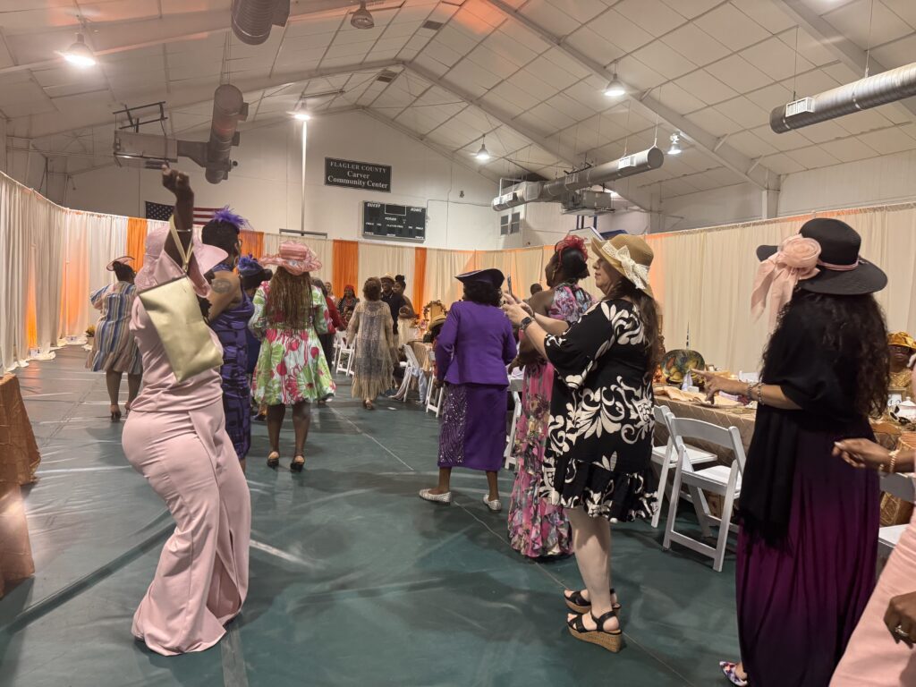 Women in colorful dresses and hats dance and mingle in a decorated indoor venue with draped curtains, tables, and chairs, suggesting a festive or formal event