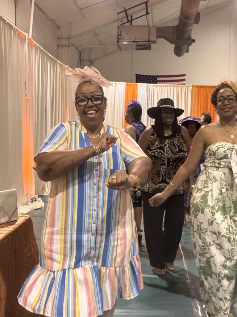 A smiling woman in a colorful striped dress dances in the foreground, while other women, some wearing hats and patterned outfits, dance behind her The scene takes place indoors with curtains and an American flag in the background