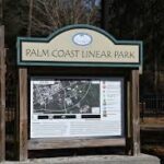 A wooden sign at the entrance of Palm Coast Linear Park displays a map and information about the park, surrounded by trees and natural scenery