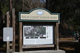 A wooden sign at the entrance of Palm Coast Linear Park displays a map and information about the park, surrounded by trees and natural scenery