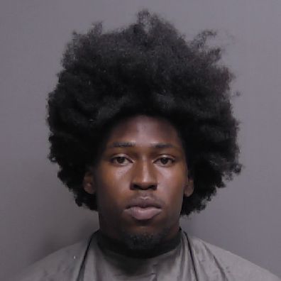 A man with a serious expression and a large afro hairstyle faces the camera against a plain gray background, wearing a dark colored garment A man with a serious expression and a large afro hairstyle faces the camera against a plain gray background, wearing a dark colored garment