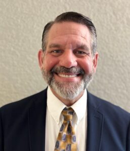 A man with short dark hair and a gray beard smiles at the camera He is wearing a navy blue suit, white shirt, and a patterned yellow tie, standing against a textured light colored wall