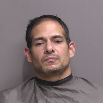A man with short dark hair and facial stubble wearing a dark colored drape sits against a plain gray background, looking directly at the camera with a neutral or slightly serious expression A man with short dark hair and facial stubble wearing a dark colored drape sits against a plain gray background, looking directly at the camera with a neutral or slightly serious expression