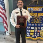 A man in a white uniform shirt and black pants stands indoors, smiling and holding a plaque Behind him are an American flag and a Kiwanis Honors banner with various patches and symbols