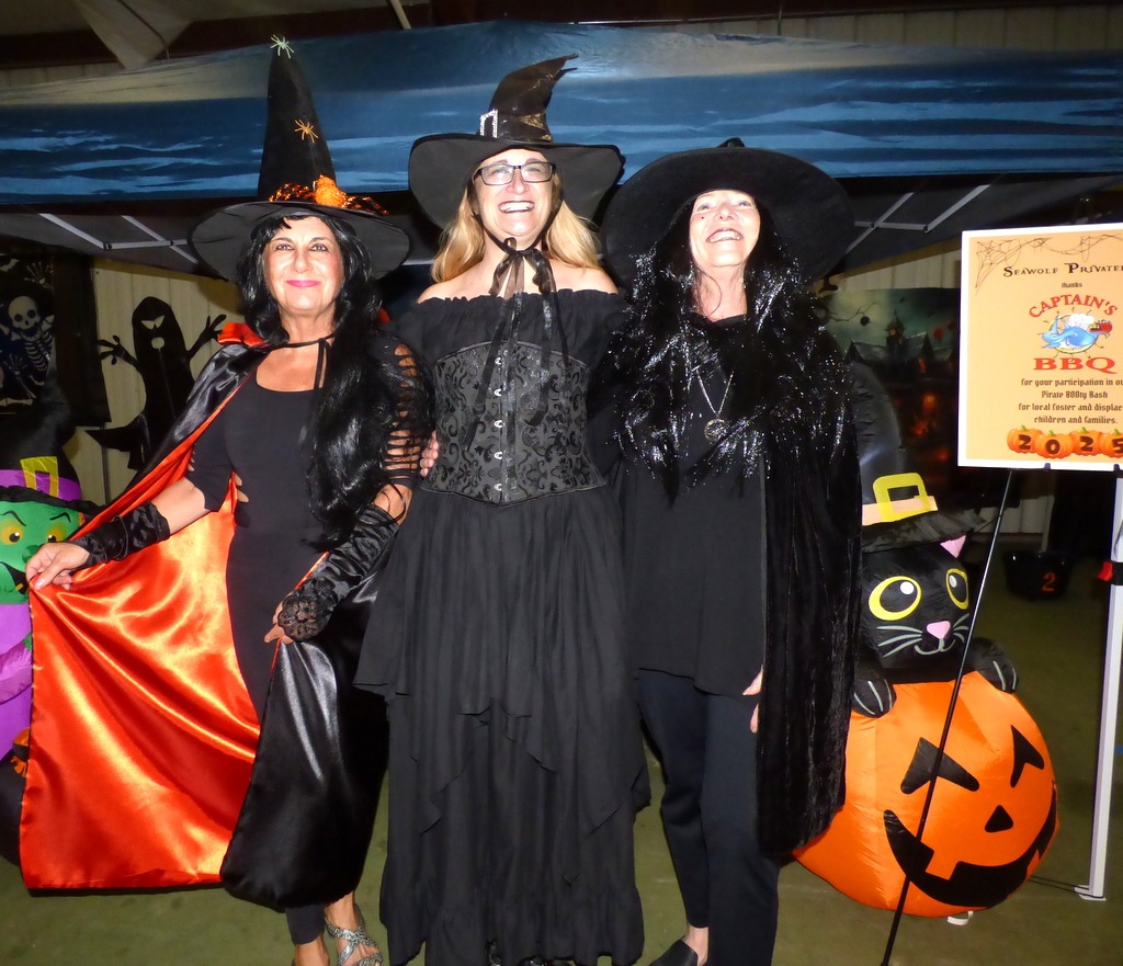 Three women dressed as witches in black costumes and hats pose and smile at a Halloween themed event, with festive decorations and a sign for a BBQ in the background