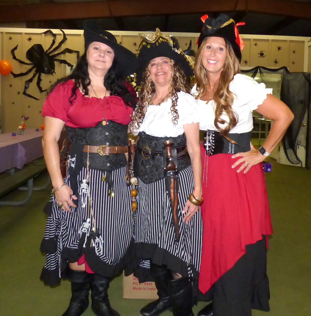 Three women dressed as pirates, wearing black hats, striped skirts, and red or white tops, stand together smiling at a Halloween themed event with spider decorations and cobwebs in the background