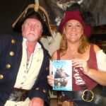 A man and woman dressed in pirate costumes smile at the camera; the woman holds up a book titled "A Place in the Pirate Realm " Shelves and a pirate flag are visible in the background