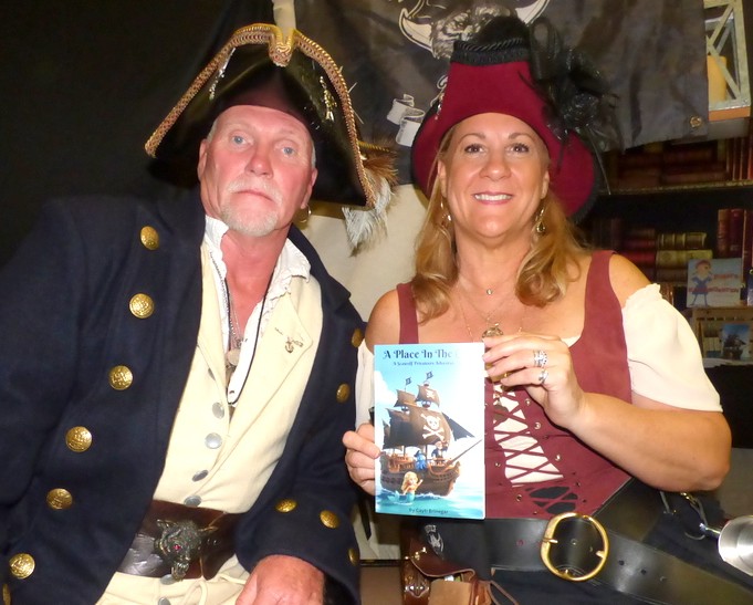A man and woman dressed in pirate costumes smile at the camera; the woman holds up a book titled "A Place in the Pirate Realm " Shelves and a pirate flag are visible in the background