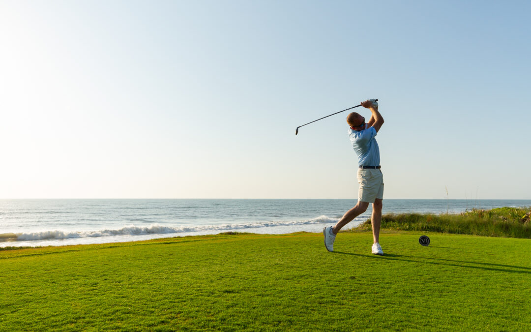 A golfer in a blue shirt and khaki shorts swings a club on a green course by the ocean under a clear blue sky, with sunlight illuminating the scene