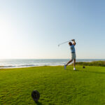 A golfer in a blue shirt and khaki shorts swings a club on a green course by the ocean under a clear blue sky, with sunlight illuminating the scene