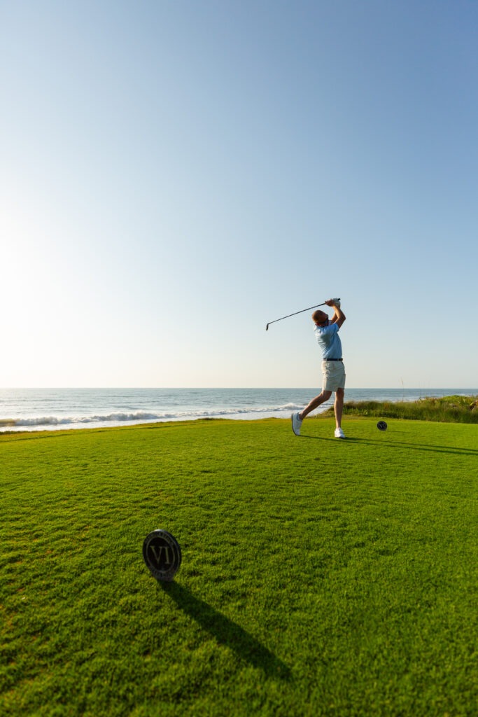 A golfer in a blue shirt and khaki shorts swings a club on a green course by the ocean under a clear blue sky, with sunlight illuminating the scene