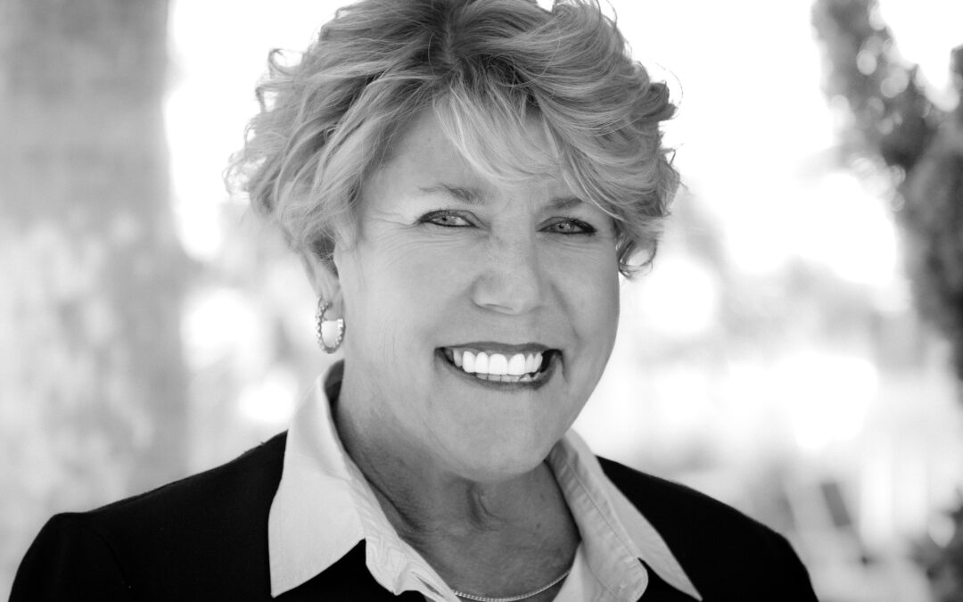 A woman with short, wavy hair smiles broadly while wearing a blazer and collared shirt The black and white photo has a blurred outdoor background