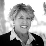 A woman with short, wavy hair smiles broadly while wearing a blazer and collared shirt The black and white photo has a blurred outdoor background