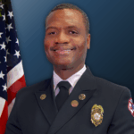 A smiling firefighter in formal uniform poses in front of a U S flag and a blue background He wears a badge and insignia on his jacket