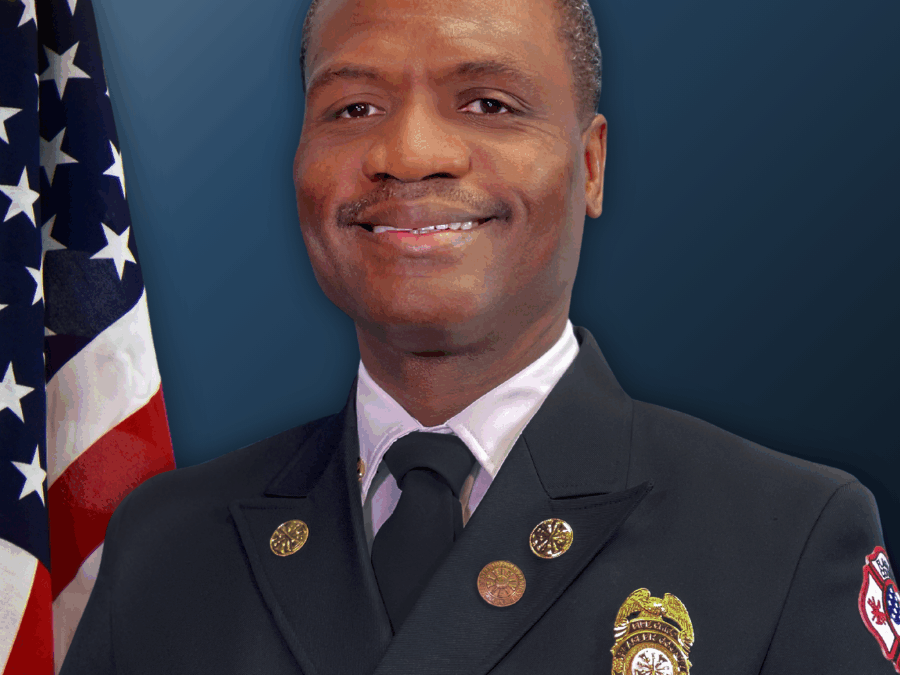 A smiling firefighter in formal uniform poses in front of a U S flag and a blue background He wears a badge and insignia on his jacket